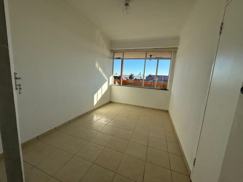 A bright, empty bedroom featuring tiled flooring, a built-in cupboard, and a large window offering an urban view.
