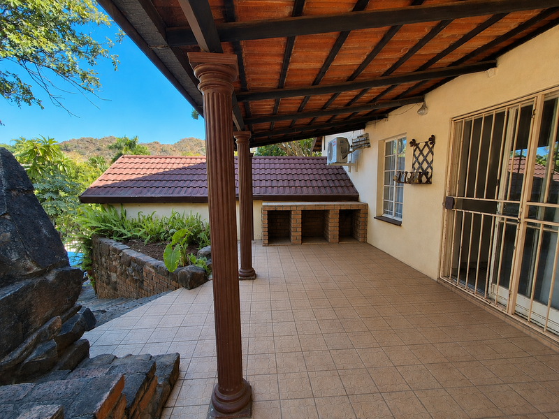 Covered patio area featuring tiled flooring, exposed timber beams, a built-in brick braai, and direct access to the garden through security gates.