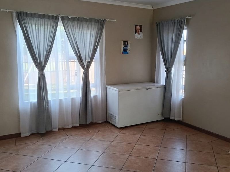 A living area featuring neutral wall tones, tiled flooring, and large windows with curtain fixtures, providing a bright and functional space.