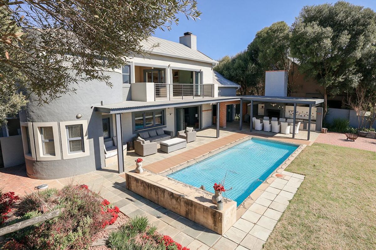 Spacious rear exterior featuring a swimming pool, covered patio with built-in braai, and an upper-level balcony overlooking the landscaped garden.
