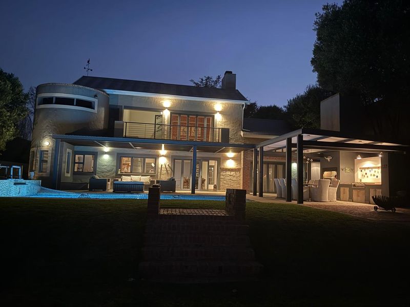 A beautifully illuminated rear exterior featuring a swimming pool, expansive covered patio, and a built-in braai area, perfect for evening outdoor entertaining.