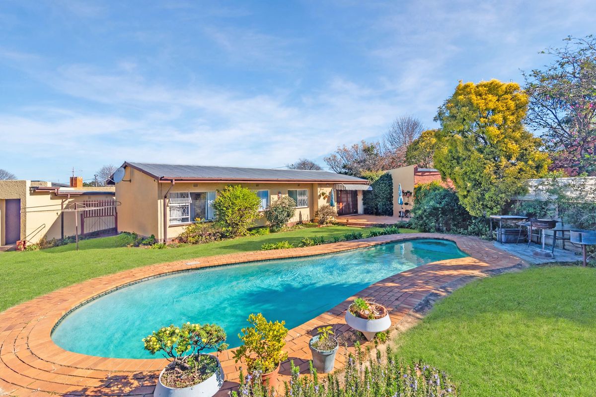 Rear view of the property featuring a swimming pool with brick coping, a well-maintained lawn, and established garden landscaping.