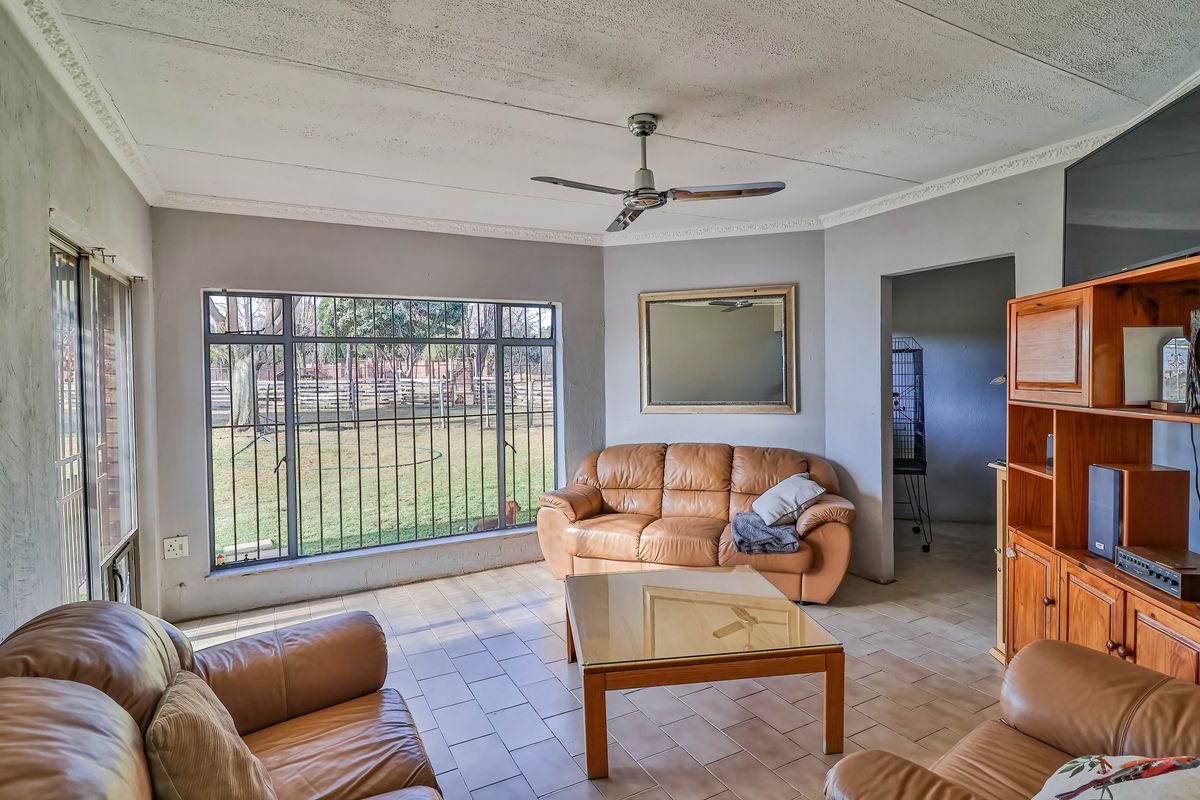 A spacious living room featuring tiled flooring, a ceiling fan, and large windows with security bars overlooking the garden.