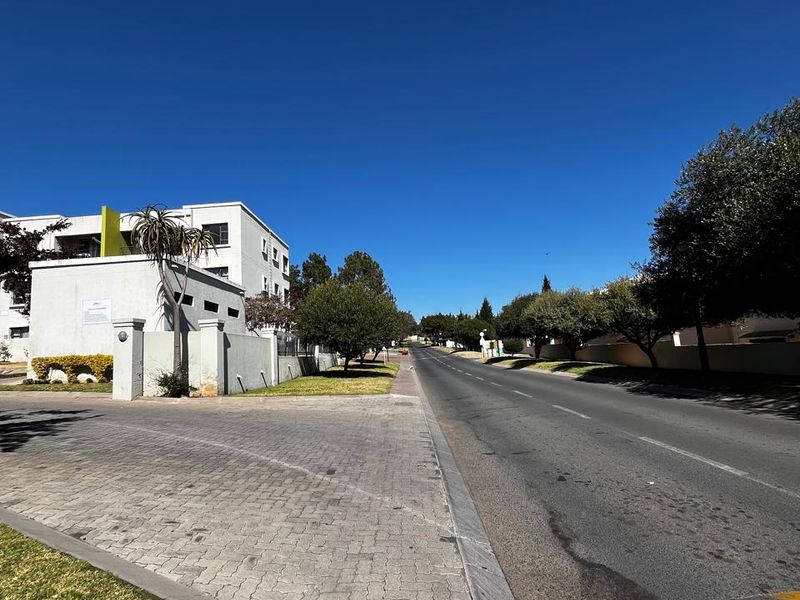 A clear street view showing the paved entrance and boundary wall of the property complex with surrounding trees and asphalt road.