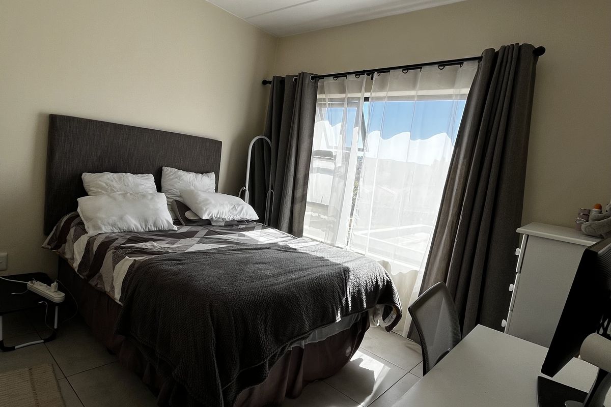 A functional bedroom featuring neutral tiled flooring, a ceiling light fixture, and large windows with curtain rails allowing for natural light.