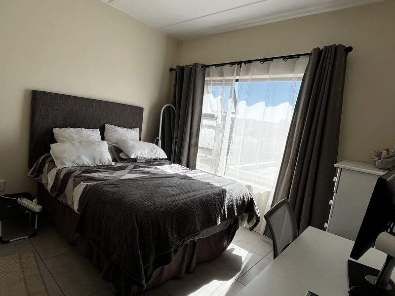 A functional bedroom featuring neutral tiled flooring, a ceiling light fixture, and large windows with curtain rails allowing for natural light.