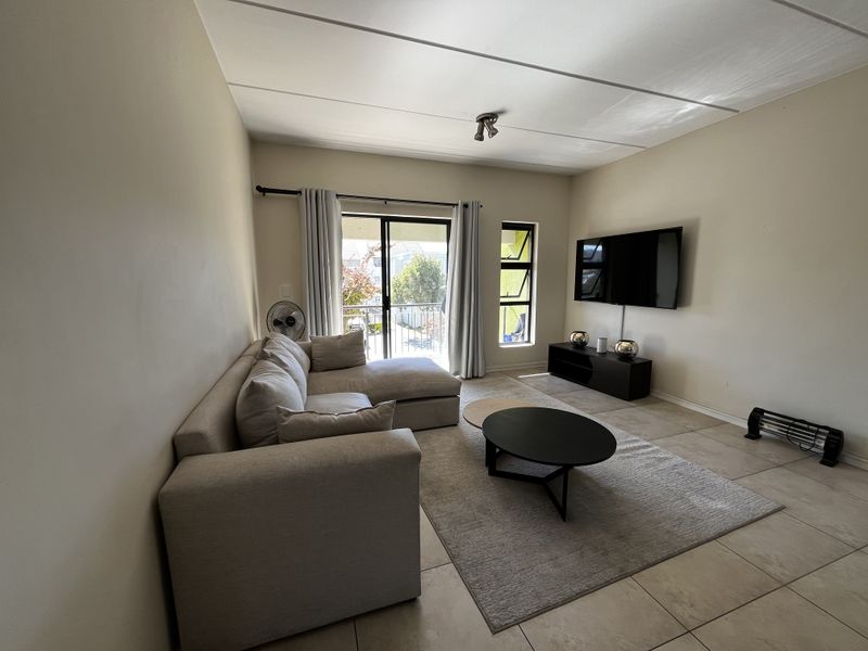 A bright living room featuring tiled flooring, a sliding glass door leading to a balcony, and a wall-mounted television setup.