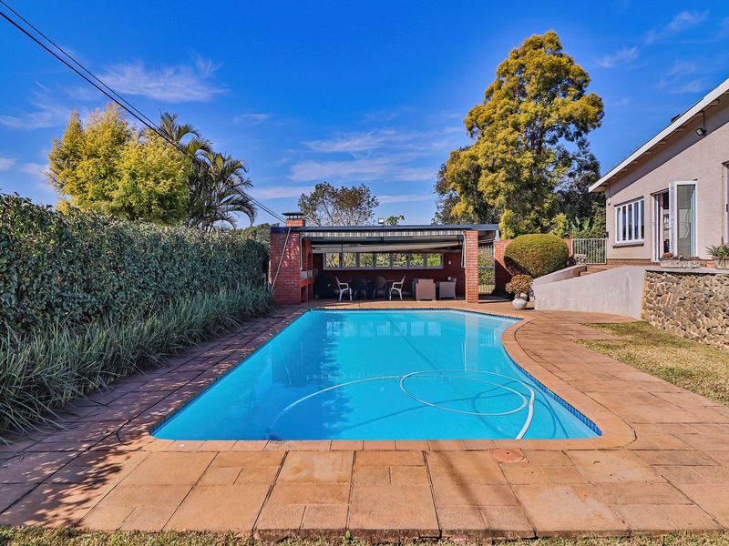 Outdoor pool area featuring a paved deck, stone-clad retaining wall, and a covered patio with a built-in brick braai.