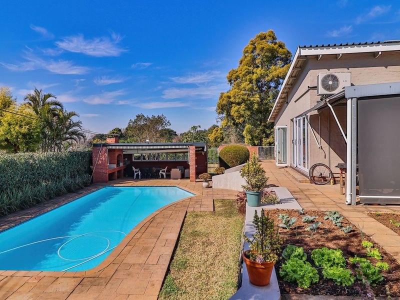 Outdoor entertainment area featuring a swimming pool, paved terrace, and a brick built-in braai, set against a landscaped garden.