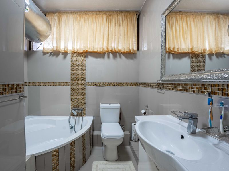 A well-maintained bathroom featuring a corner bathtub, pedestal basin, and modern tiled finishes with a decorative mosaic border.