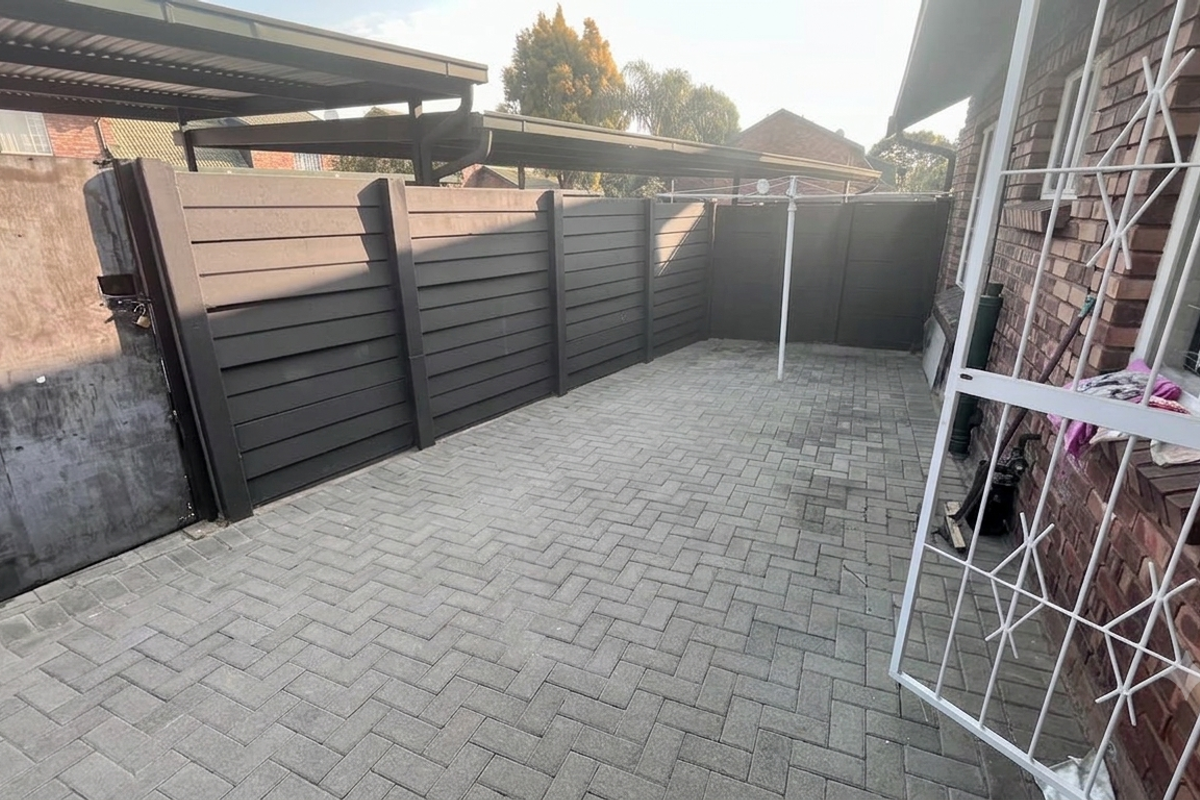 A paved rear courtyard area featuring perimeter fencing, a security gate, and access to a covered carport structure.