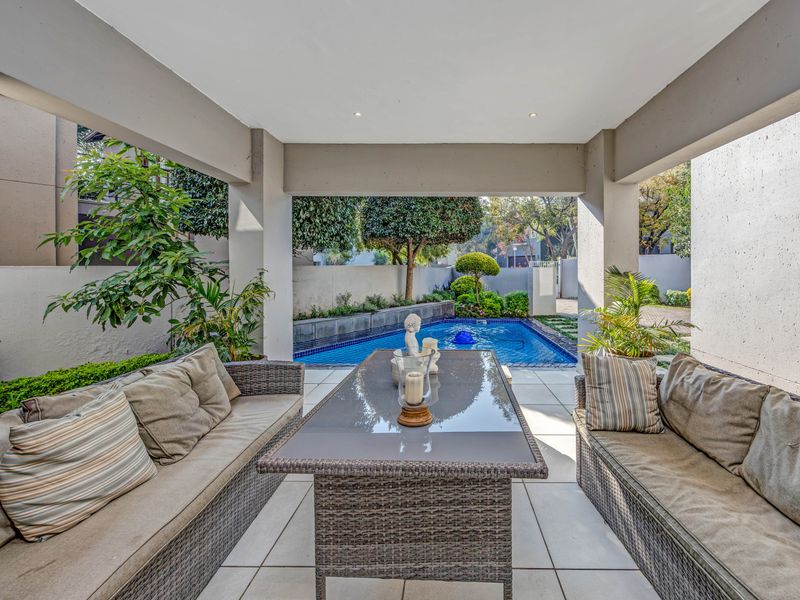 A covered outdoor patio featuring tiled flooring and recessed lighting, overlooking a private swimming pool and landscaped garden area.