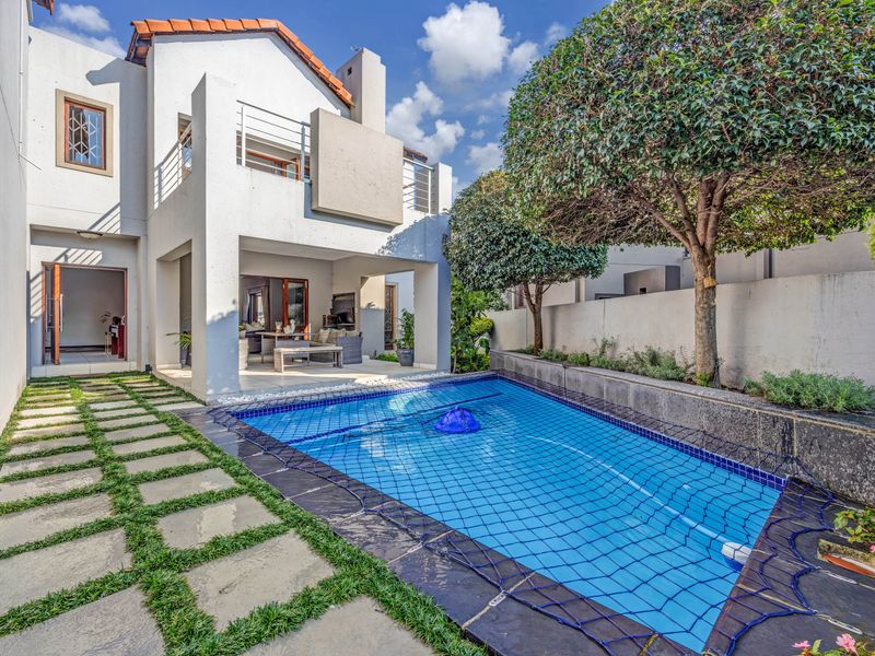 A well-maintained rear garden area featuring a swimming pool with safety net, stone-paved walkway, and a covered patio attached to the main house structure.