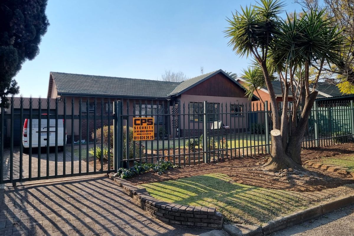 Front elevation of a residential house featuring a tiled roof, paved driveway, and secure metal perimeter fencing with a gated entrance.