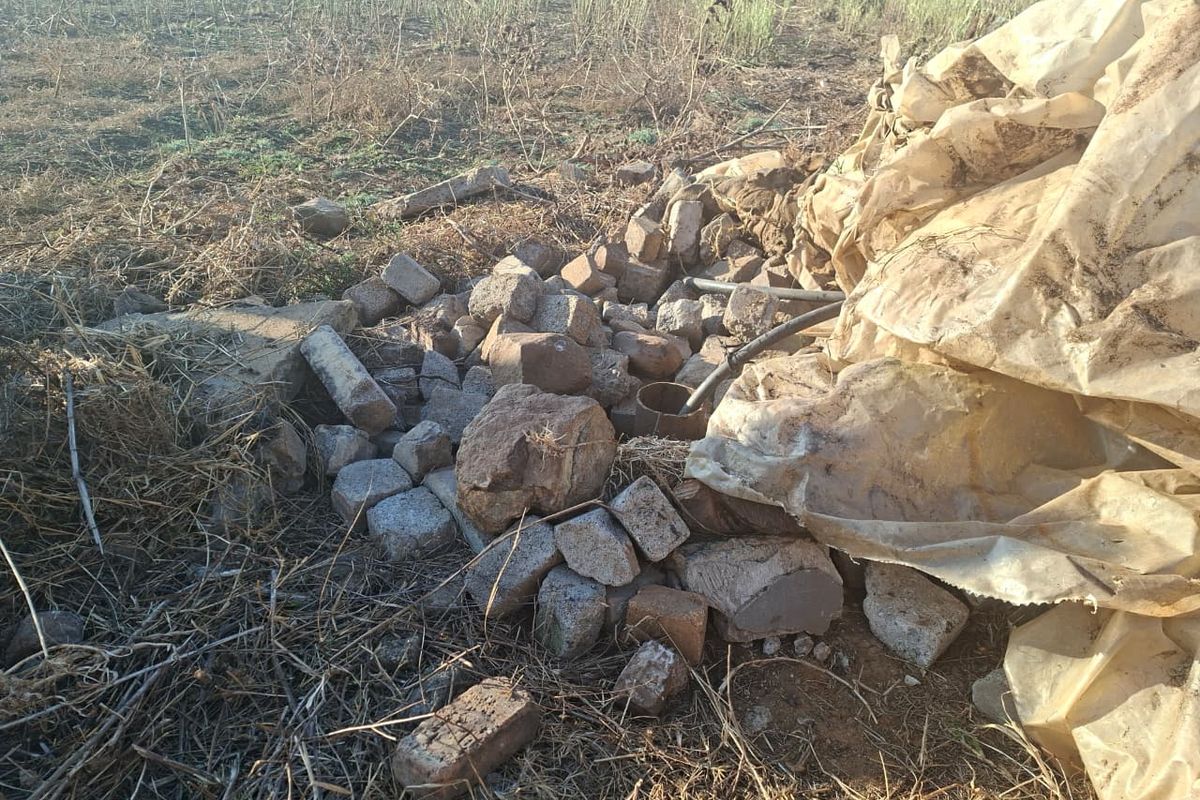 An unmaintained outdoor area featuring a borehole casing surrounded by loose stone debris and discarded materials on the small holding.