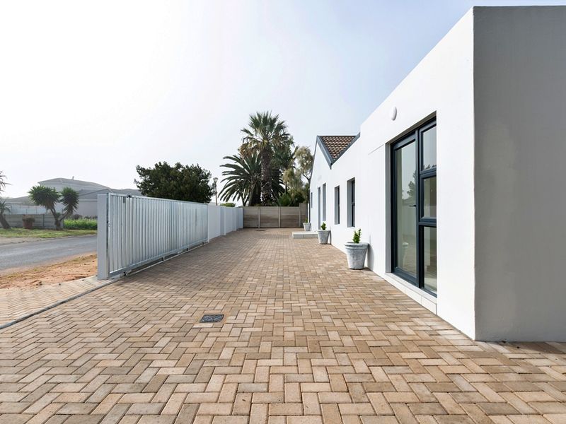 A clean exterior view of the property featuring a paved driveway, secure sliding gate, and modern white facade with dark aluminium window frames.
