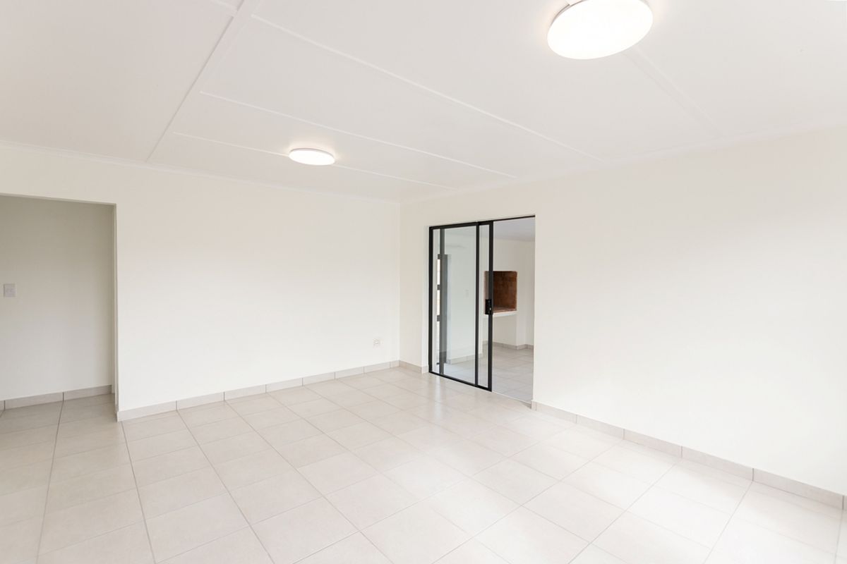 A bright, empty living area featuring neutral tiled flooring, clean white walls, and a modern black-framed glass door leading to an adjacent space.