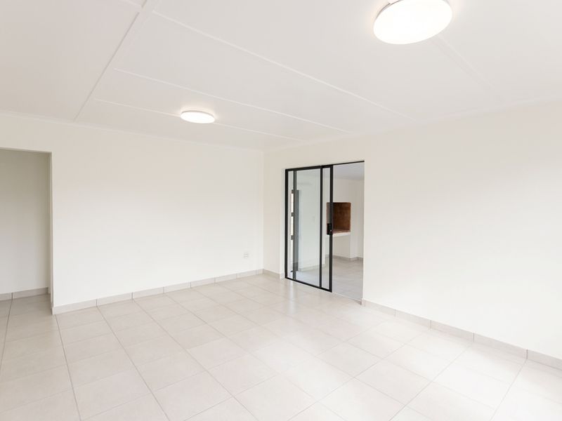 A bright, empty living area featuring neutral tiled flooring, clean white walls, and a modern black-framed glass door leading to an adjacent space.