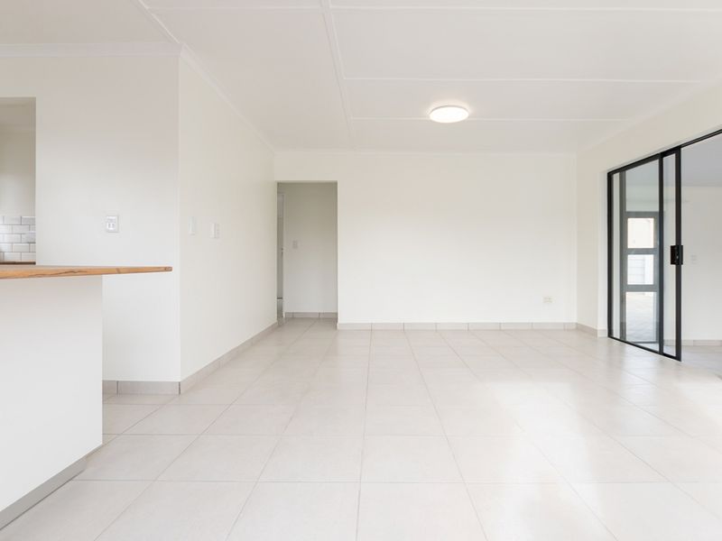 A bright, open-plan living area featuring neutral tiled flooring, clean white walls, and a seamless transition to the kitchen and outdoor space.