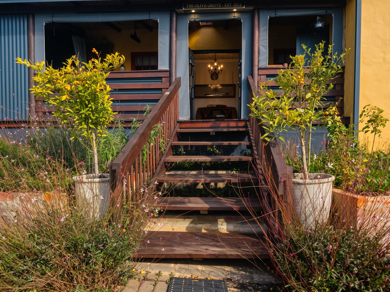 A charming front entrance featuring a timber staircase leading to a covered veranda with corrugated metal accents and established garden landscaping.