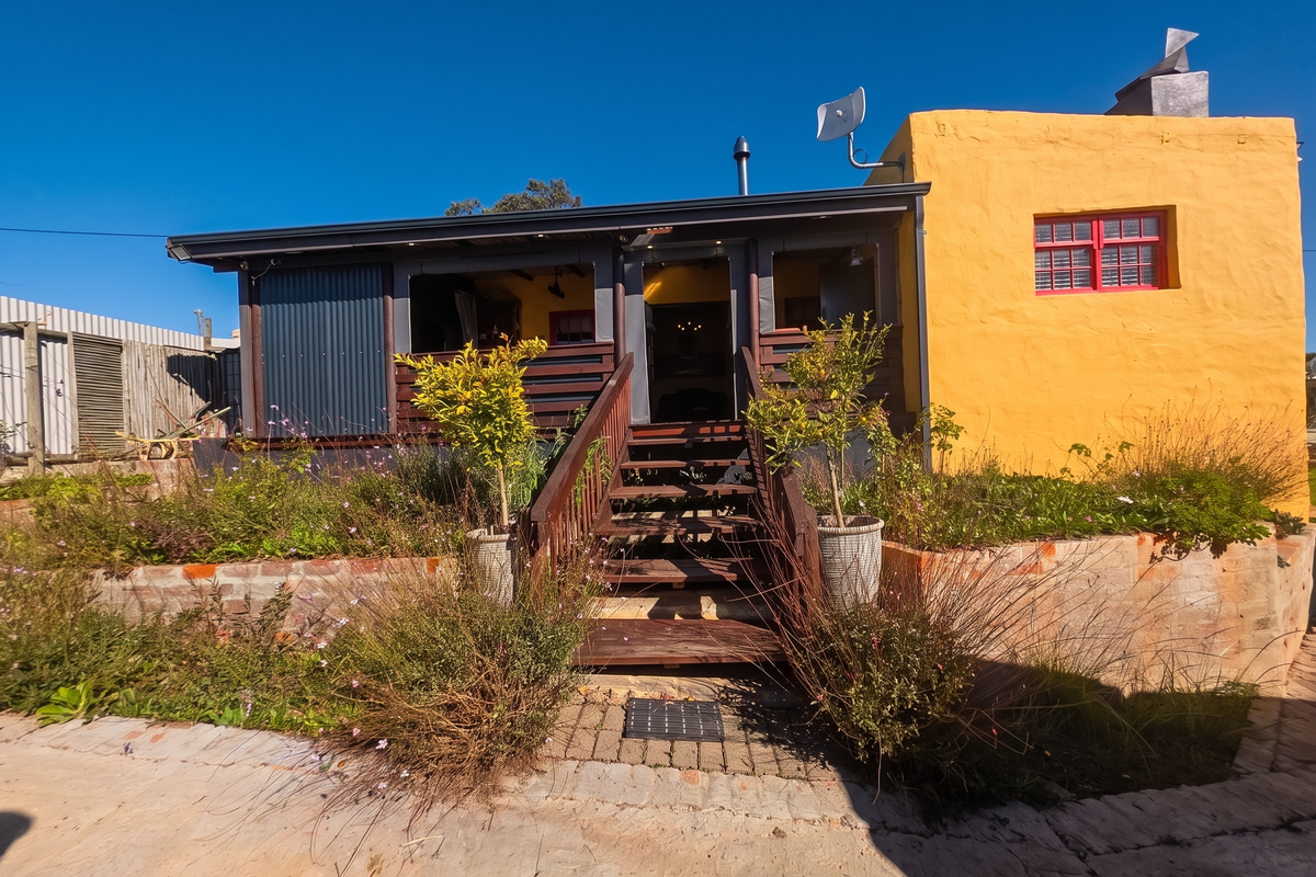 Rear exterior view featuring a raised timber deck with staircase, corrugated metal accents, and a vibrant yellow rendered wall with brick retaining garden beds.