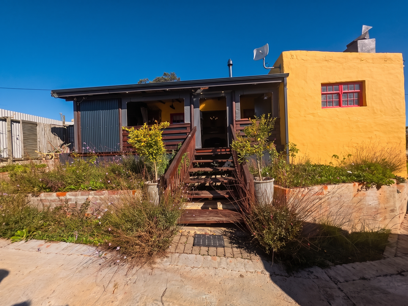 Rear exterior view featuring a raised timber deck with staircase, corrugated metal accents, and a vibrant yellow rendered wall with brick retaining garden beds.