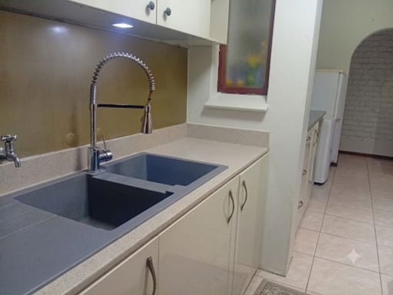 A clean, functional kitchen area featuring a modern double basin sink with a gooseneck faucet, ample built-in cabinetry, and neutral tiled flooring.
