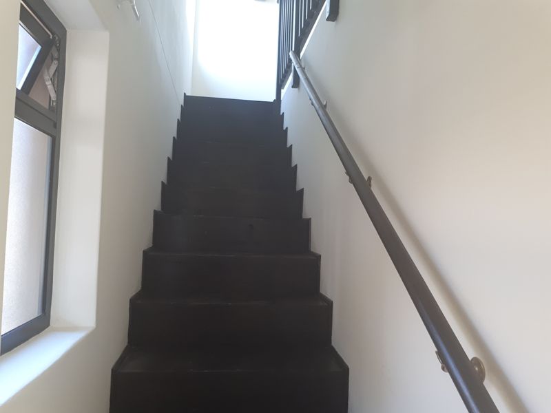 A simple interior staircase featuring dark-painted treads and a wall-mounted handrail, illuminated by natural light from an adjacent window.