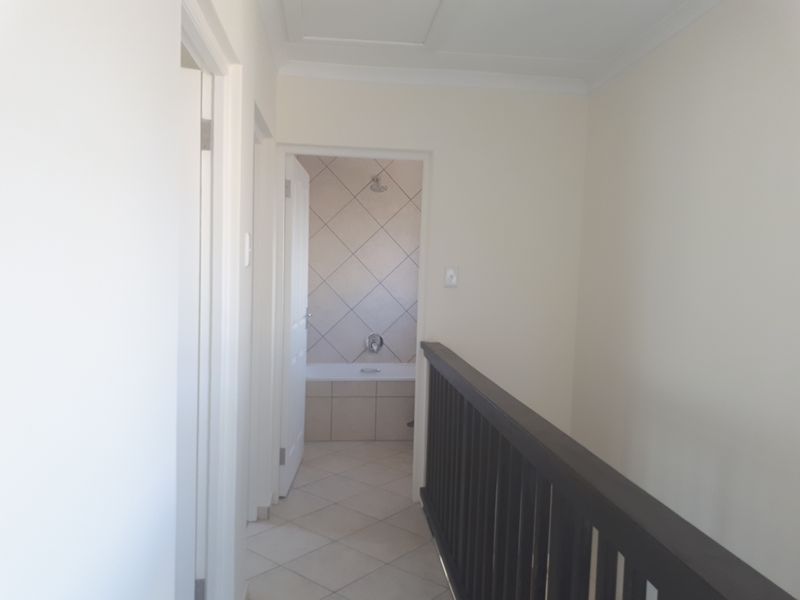 A bright, neutral hallway featuring tiled flooring and a dark timber balustrade, providing access to adjacent rooms including a bathroom.