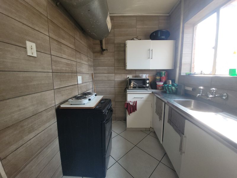 A compact kitchen featuring tiled walls, built-in cabinetry, a stainless steel sink, and exposed ventilation ducting, requiring modernization to improve functionality and aesthetic appeal.