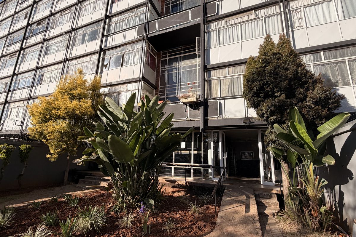 Front exterior view of a multi-story apartment building featuring a paved entrance walkway and established garden beds.