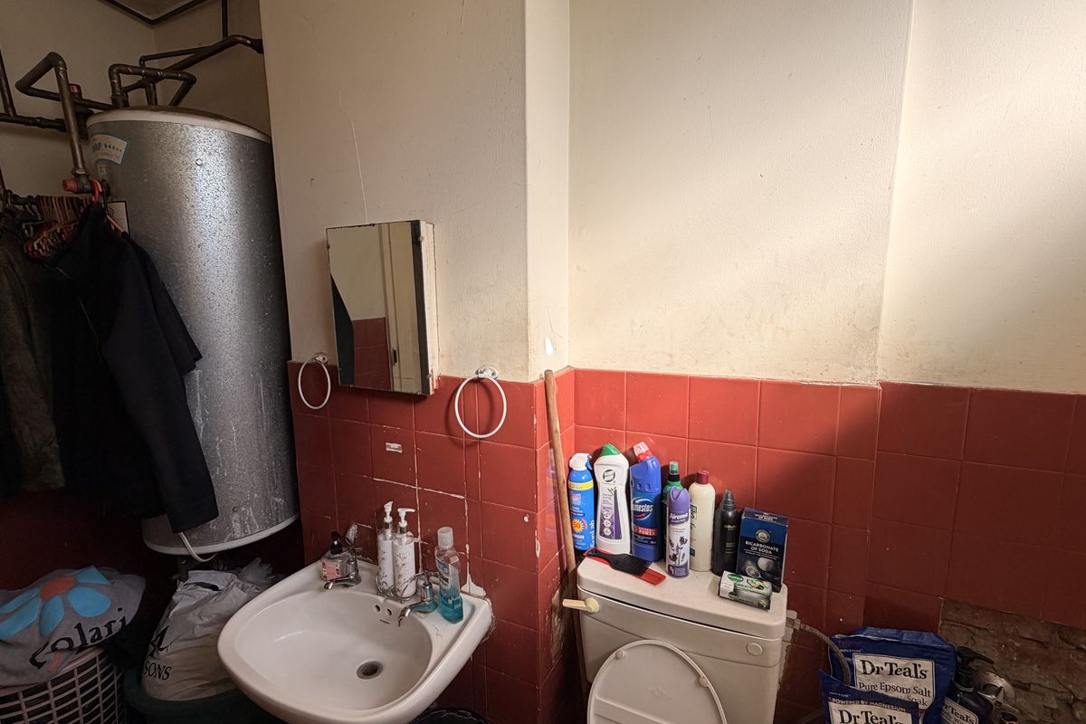 A dated bathroom featuring a wall-mounted electric water heater, ceramic washbasin, and toilet with red tiled splashbacks requiring renovation and cosmetic updates.