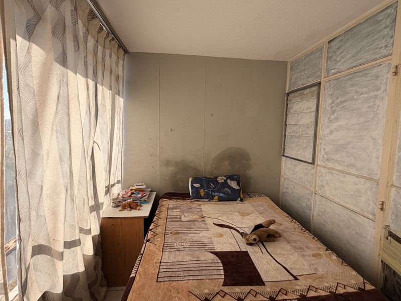 A bedroom featuring built-in cupboards and a large window, currently showing signs of wall water damage and dated finishes requiring maintenance.