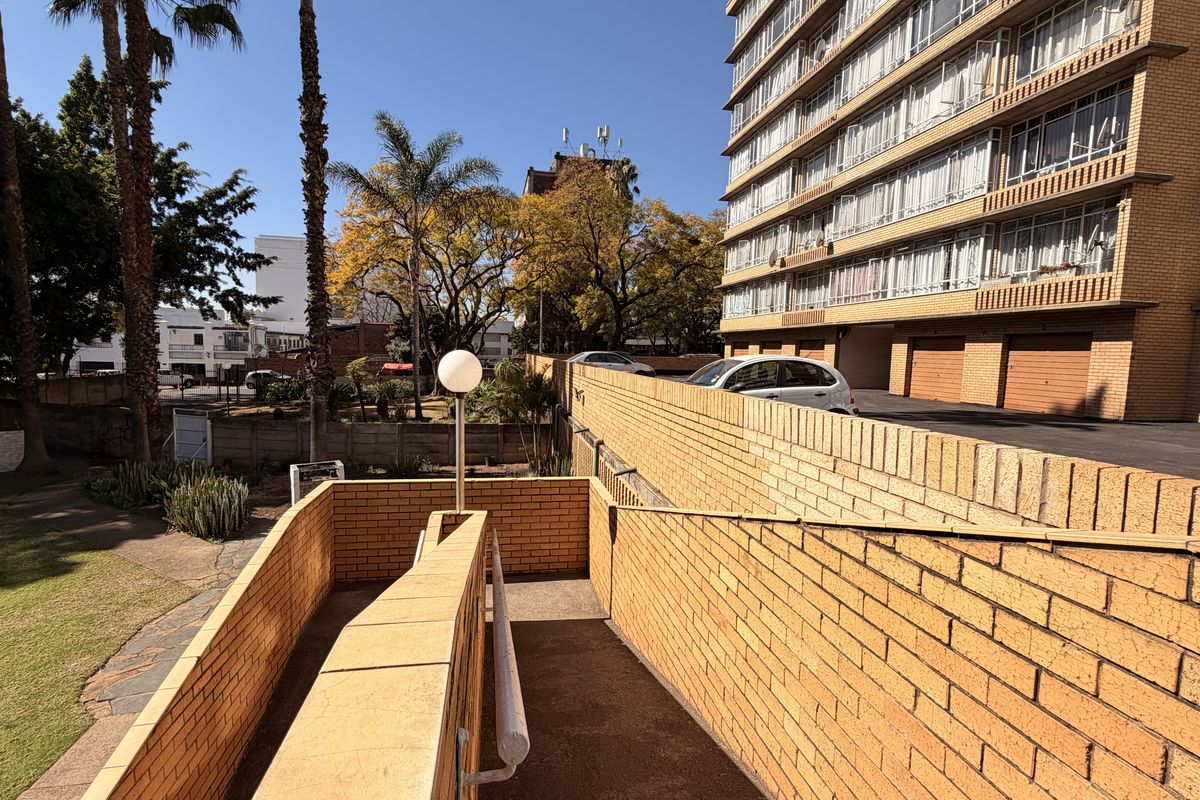 Exterior view of an apartment complex featuring a brick retaining wall, a concrete access ramp with a handrail, and landscaped garden areas.