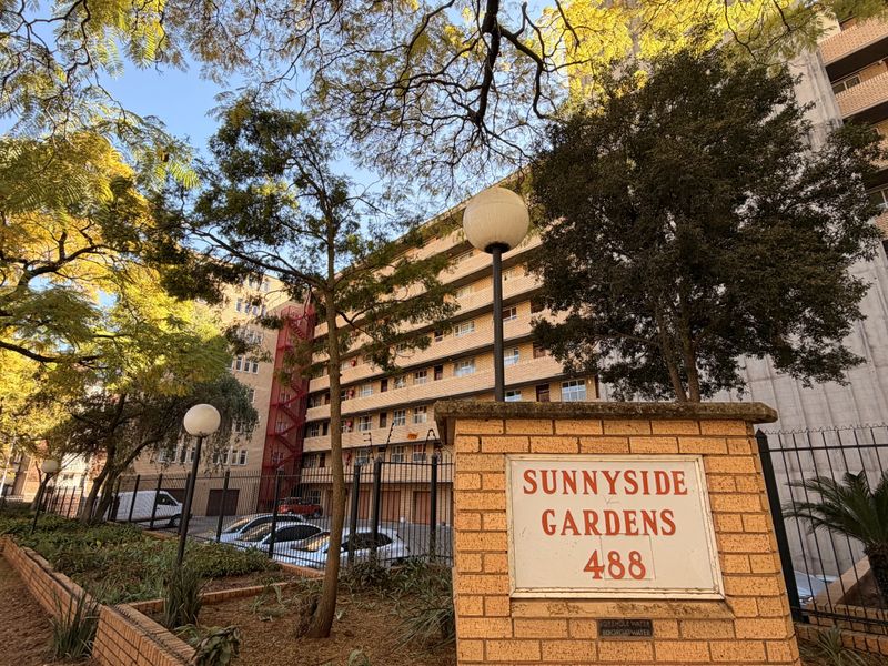 Street-level view of the Sunnyside Gardens apartment complex, featuring brick facade, security fencing, and established trees.