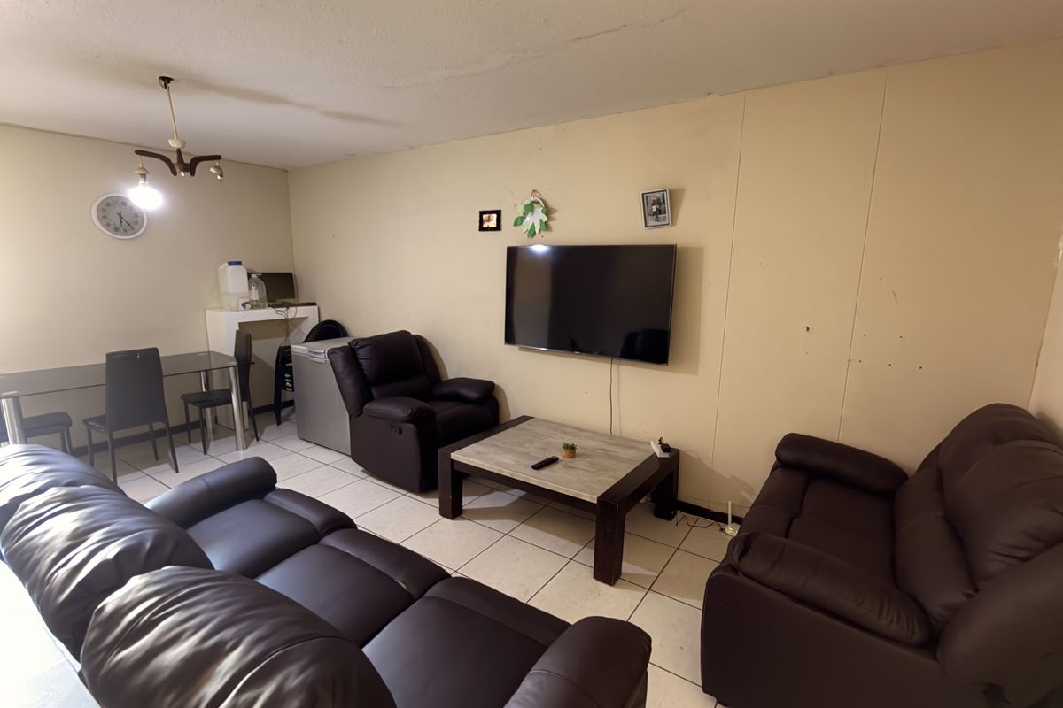 A living room area featuring tiled flooring and wall-mounted television, showing signs of wear on the walls and ceiling requiring cosmetic attention.