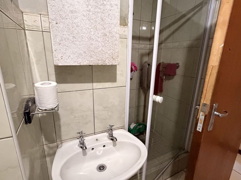 A functional bathroom featuring a pedestal basin, shower enclosure, and tiled finishes, currently showing signs of wear on the door frame and cabinetry.