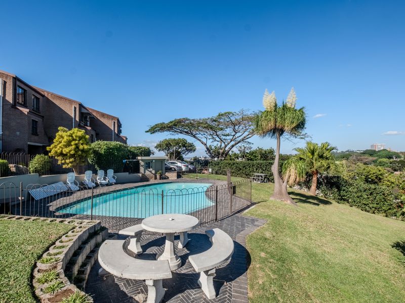 A well-maintained outdoor pool area featuring a swimming pool, paved patio space, and landscaped garden surroundings within a townhouse complex.