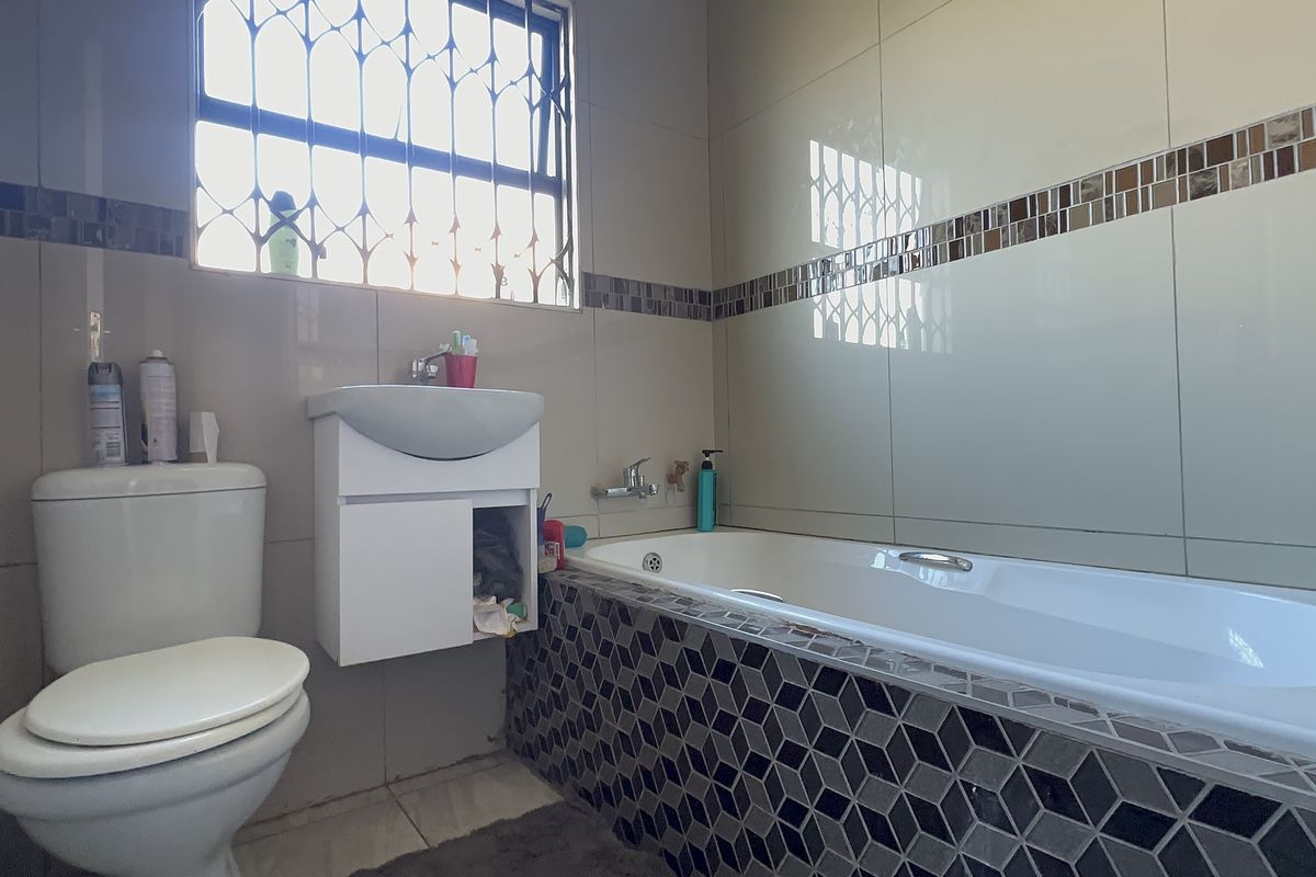 A functional bathroom featuring a bathtub with mosaic tiling, a wall-mounted vanity, and a toilet, finished with neutral wall tiles and security window bars.