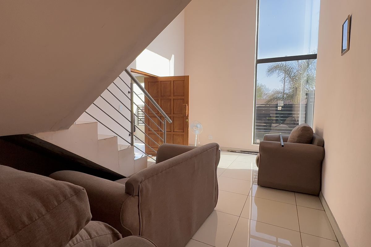Bright living area featuring high ceilings, large floor-to-ceiling windows, tiled flooring, and a modern staircase with a stainless steel balustrade.
