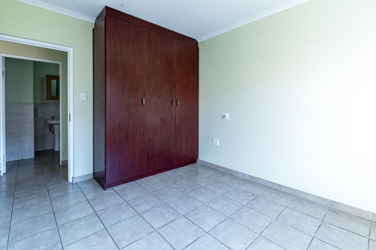 A bright, empty bedroom featuring tiled flooring and a large built-in wooden wardrobe, with an adjoining bathroom visible through the doorway.