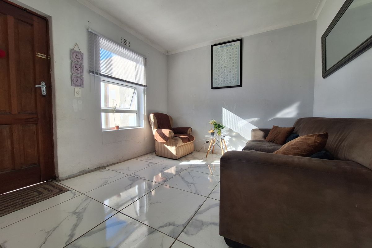 A bright living room featuring large marble-look floor tiles, a timber entrance door, and natural light through an aluminium-framed window.