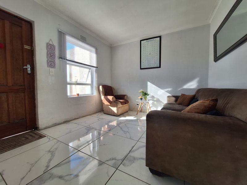 A bright living room featuring large marble-look floor tiles, a timber entrance door, and natural light through an aluminium-framed window.