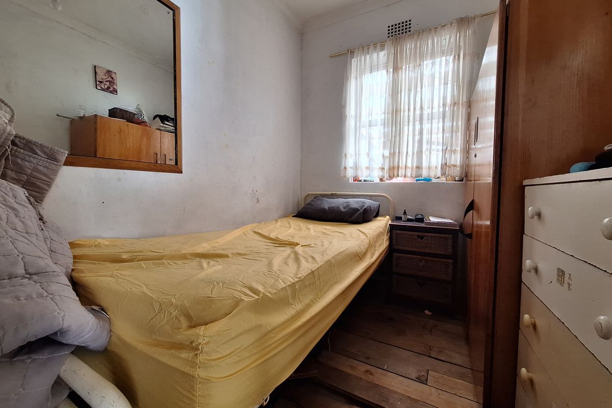 A compact bedroom featuring timber flooring, a built-in wardrobe, and a wall-mounted mirror, requiring cosmetic updates to improve its overall condition.