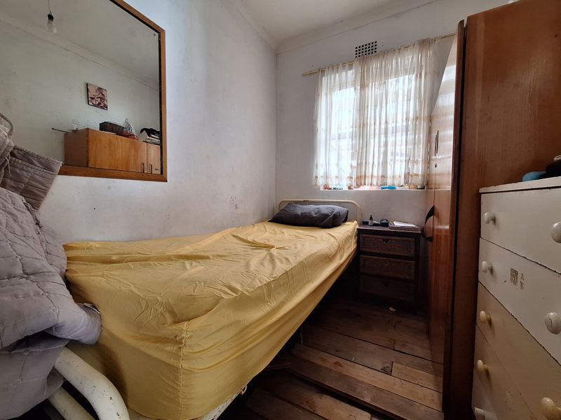 A compact bedroom featuring timber flooring, a built-in wardrobe, and a wall-mounted mirror, requiring cosmetic updates to improve its overall condition.