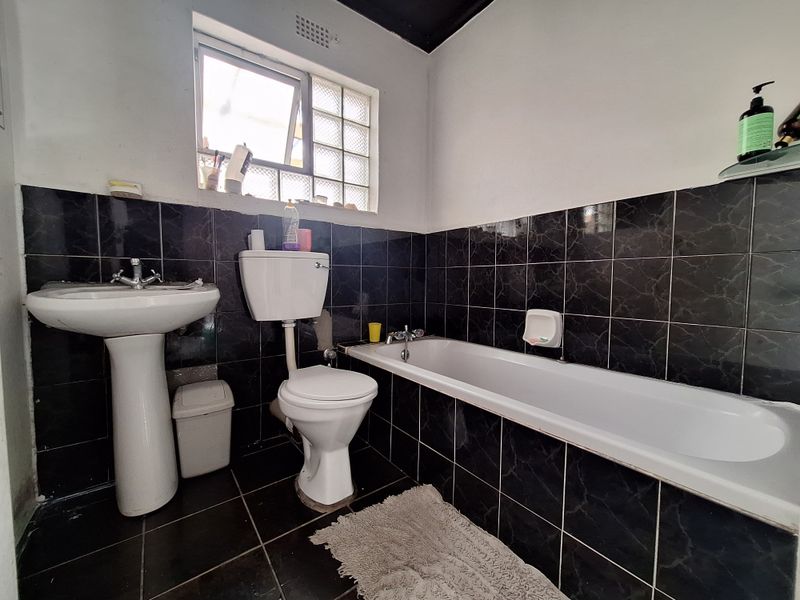 A functional bathroom featuring a pedestal basin, toilet, and bathtub with dark marble-effect wall and floor tiling and a glass block window.