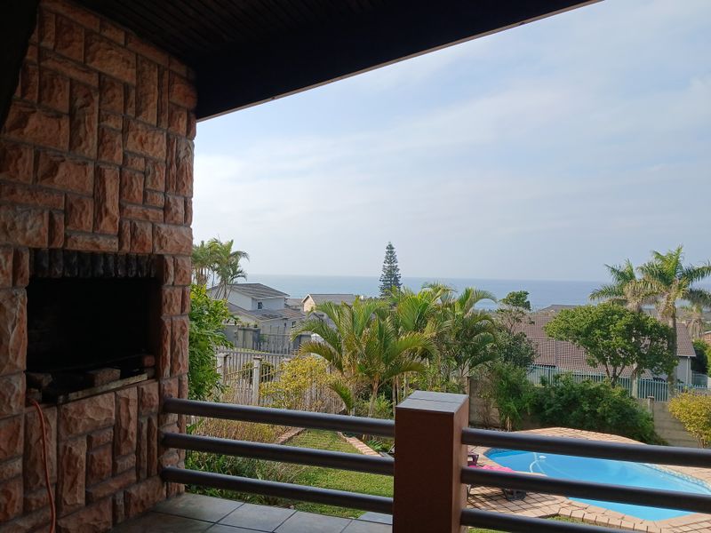 Covered patio featuring a built-in braai, stone cladding, and metal railings, overlooking the garden, swimming pool, and distant ocean view.
