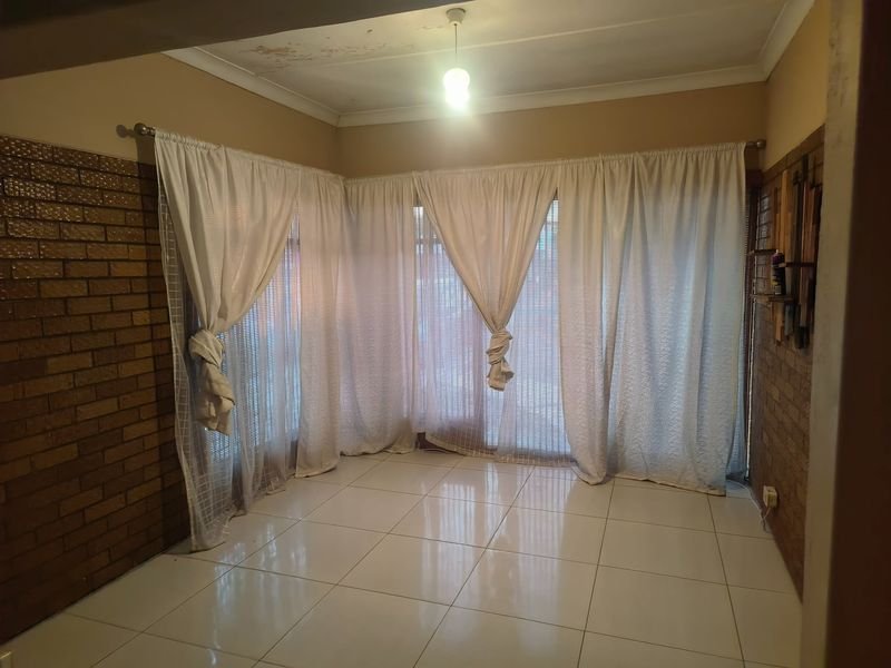An empty living area featuring exposed brick walls, tiled flooring, and large windows with curtain tracks, showing signs of ceiling water damage.