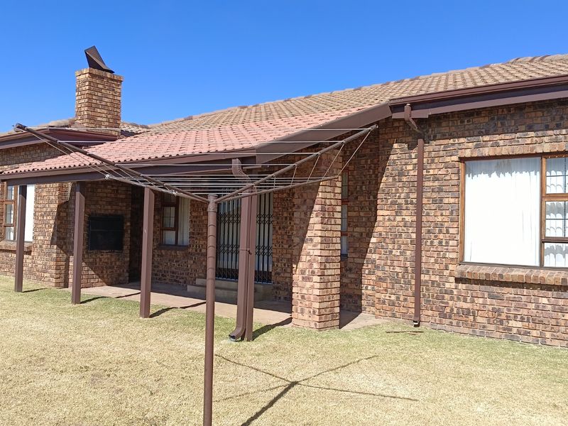 Rear view of a face-brick house featuring a covered patio with a built-in braai, tiled roof, and a well-maintained lawn.