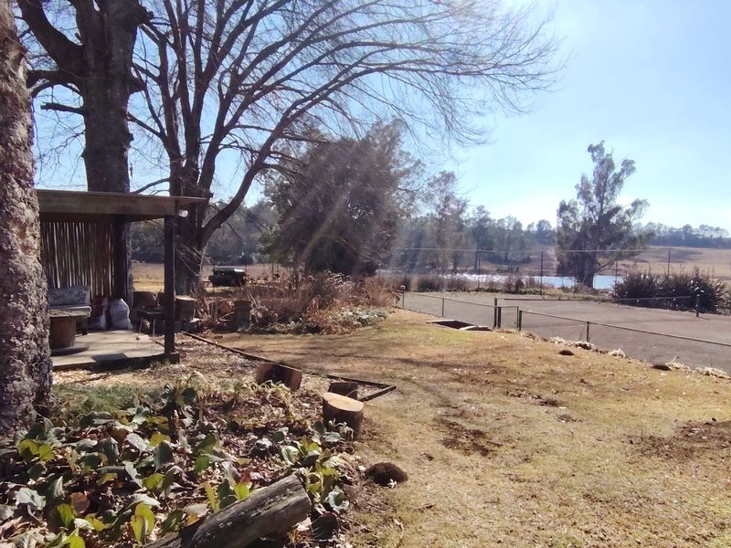 A spacious garden area featuring a covered patio structure, mature trees, and perimeter fencing with views of the surrounding landscape.